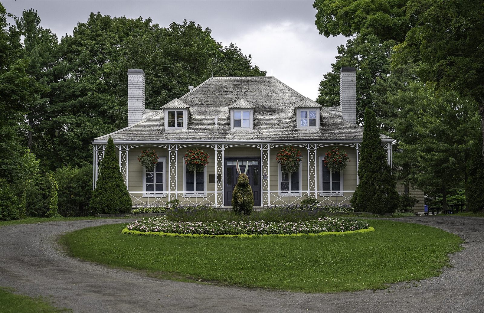 A wide house seen straight on from the far side of a gravel circular driveway. The drive loops around an island of mown grass holding a round flower bed and a rabbit topiary. A white lattice verandah runs the full width of the house, hung with baskets of red flowers, and three dormer windows and two white chimneys break the shingled roof.