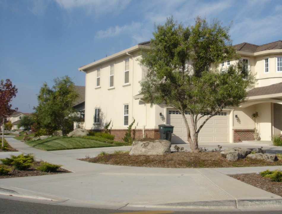 A two-storey cream stucco house standing on a street corner in bright sunshine. One elevation runs away down the side street to the left while the garage front faces the street in the foreground. A concrete sidewalk wraps around the corner with a ramp at the crossing, and the planting is gravel, low junipers and boulders rather than lawn.