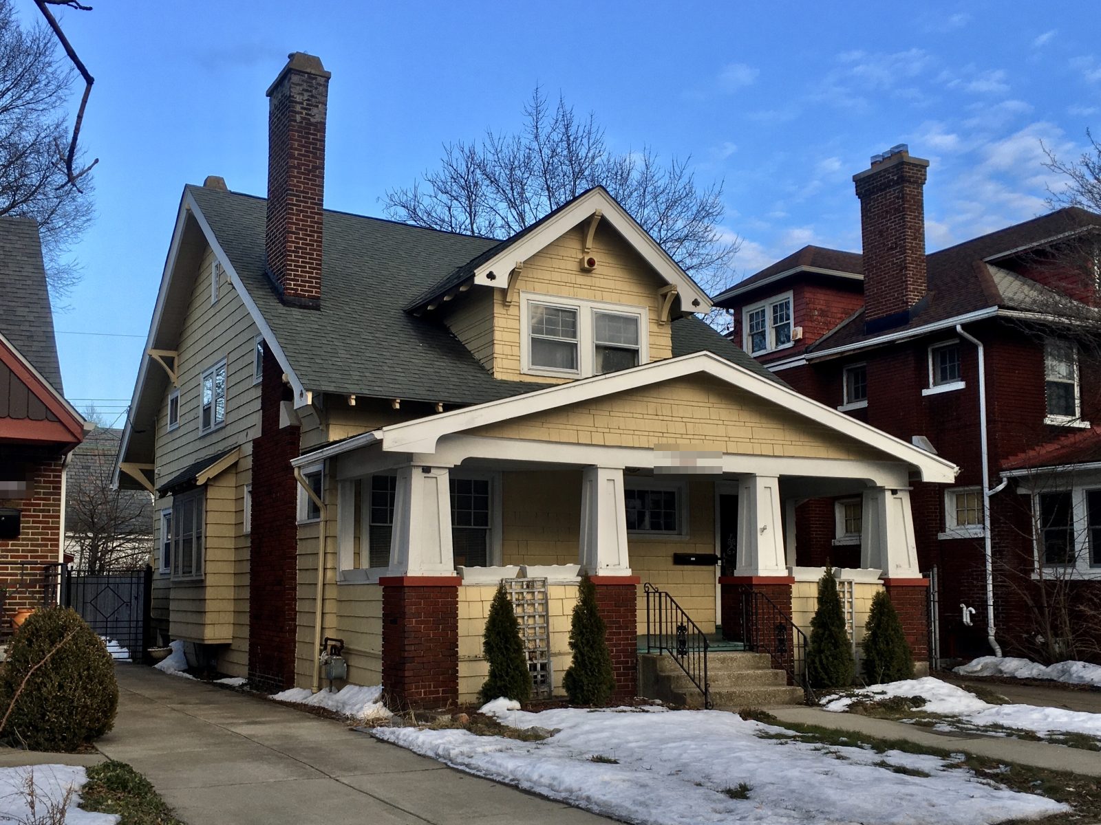 A craftsman bungalow on a narrow city lot, with a deep covered front porch on tapered pillars over brick piers, a short front lawn and neighbouring houses close on both sides.