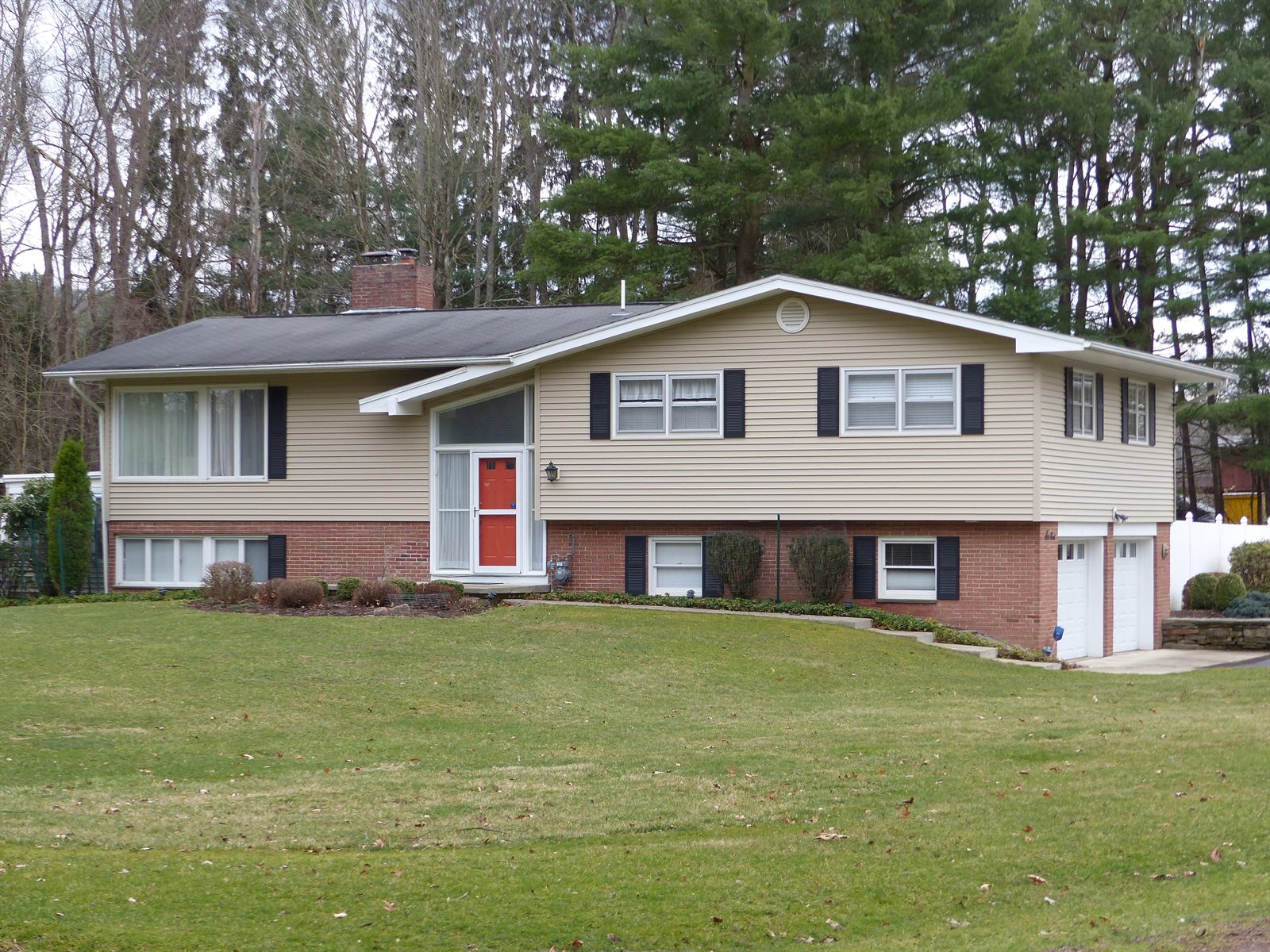 A split-level house photographed from the street on an overcast day. Tan vinyl siding above, red brick below, and a roofline that steps down from right to left. A red front door sits in a tall glazed entry bay in the middle of the facade, a few steps up from the walk. Two garage doors are set into the brick lower level at the right-hand end, reached by a driveway coming in from the right. A wide lawn rises gently from the street to the house.