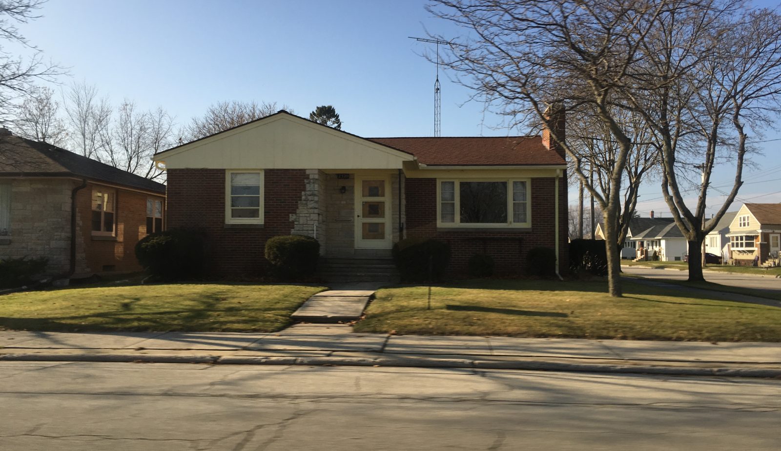 A low single-storey brick ranch house seen straight on from the street, wide across the frontage with a short front lawn and a straight walk to a small stoop. No covered porch.